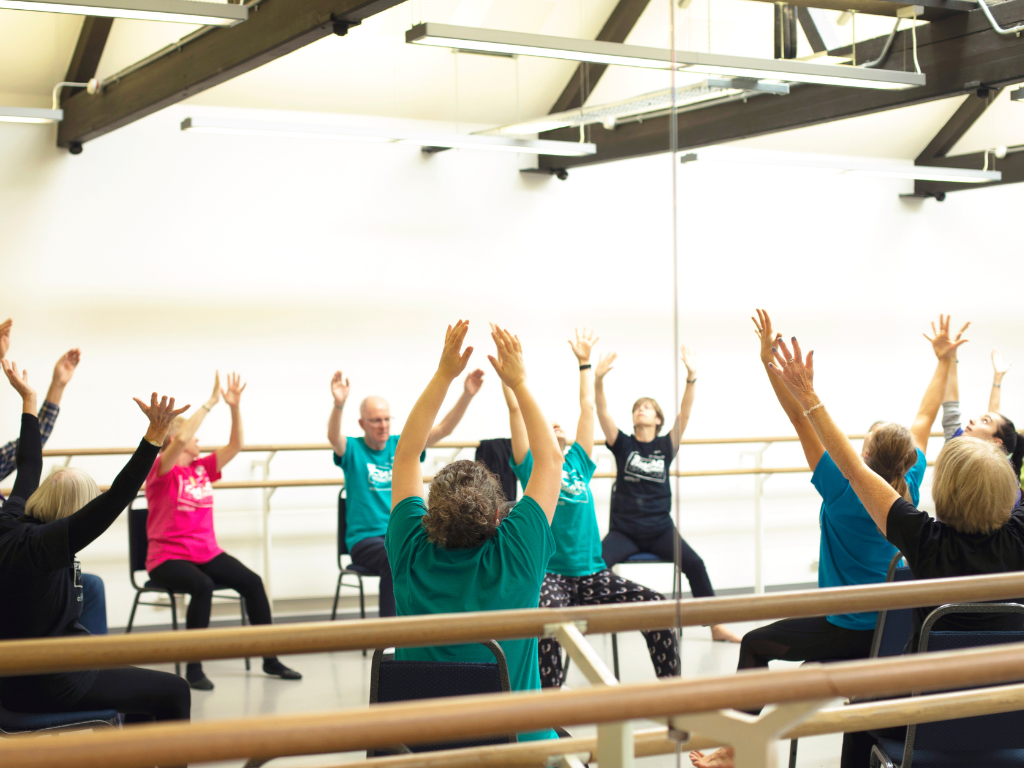 Dance for Parkinson’s Dance for Parkinson's group sitting on chairs with their hands raised in the air in a brightly lit dance studio.