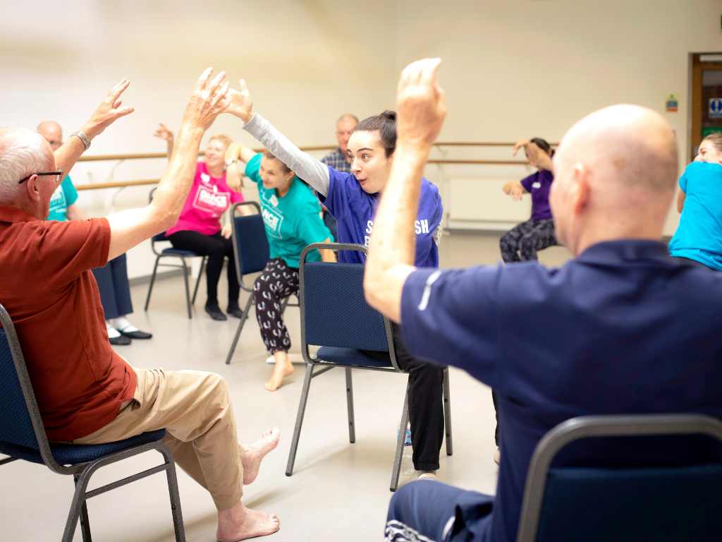 Dance for Parkinson’s Dance for Parkinson's dancers sitting on chairs with their hands raised in the air during a Dance for Parkinson's class.
