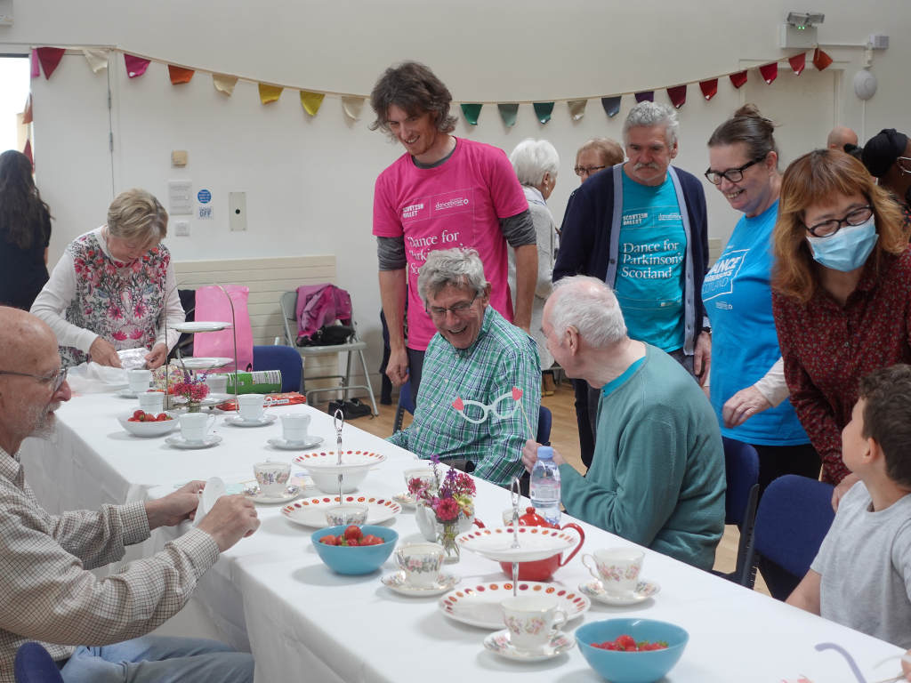 Dance for Parkinson’s tea party Dance for Parkinson's dancers siting around a table with teapots and cake stands on it. Some members are standing and smiling while others remain seated. Bunting decorates the background on the wall.