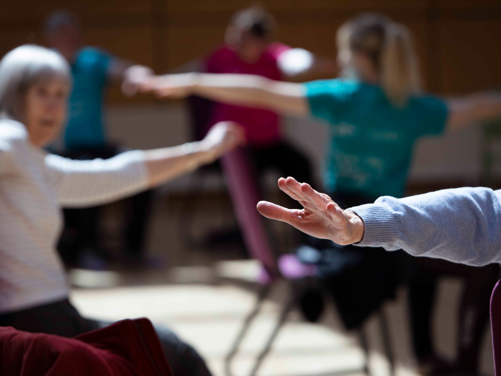 Dance for Parkinson’s A blurred image of seated dancers with one arm reaching back which remains in focus.