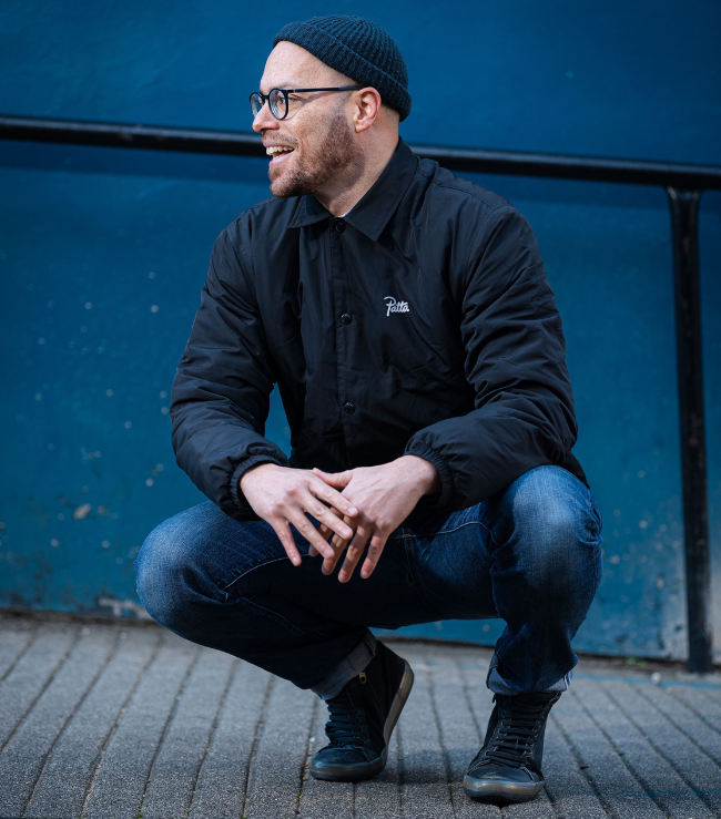 Photograph of Tony Mills squatting down in front of a blue-painted wall, and smiling off to the side. Tony wears jeans, a black jacket and a black beanie.