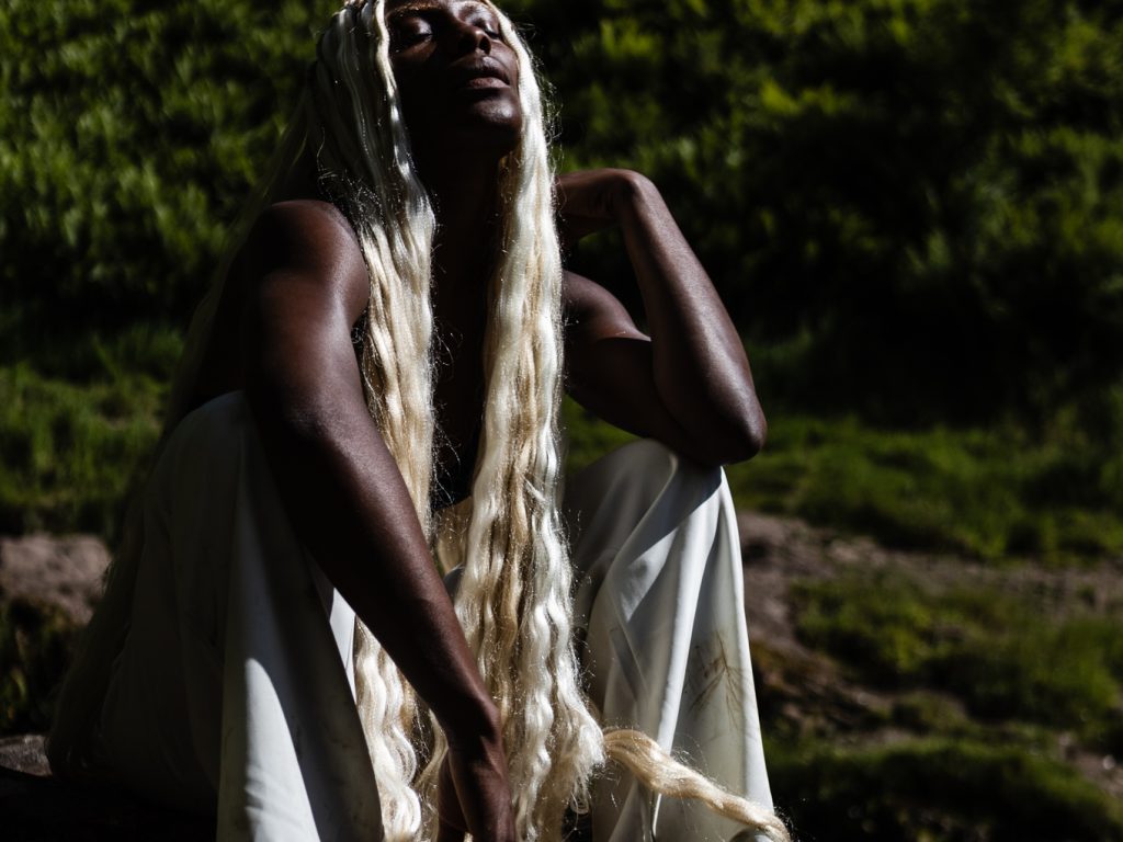 Divine Tasinda A Black woman with long, blonde hair sits on a rock. Hair eyes are closed, and she tilts her face towards the sun.