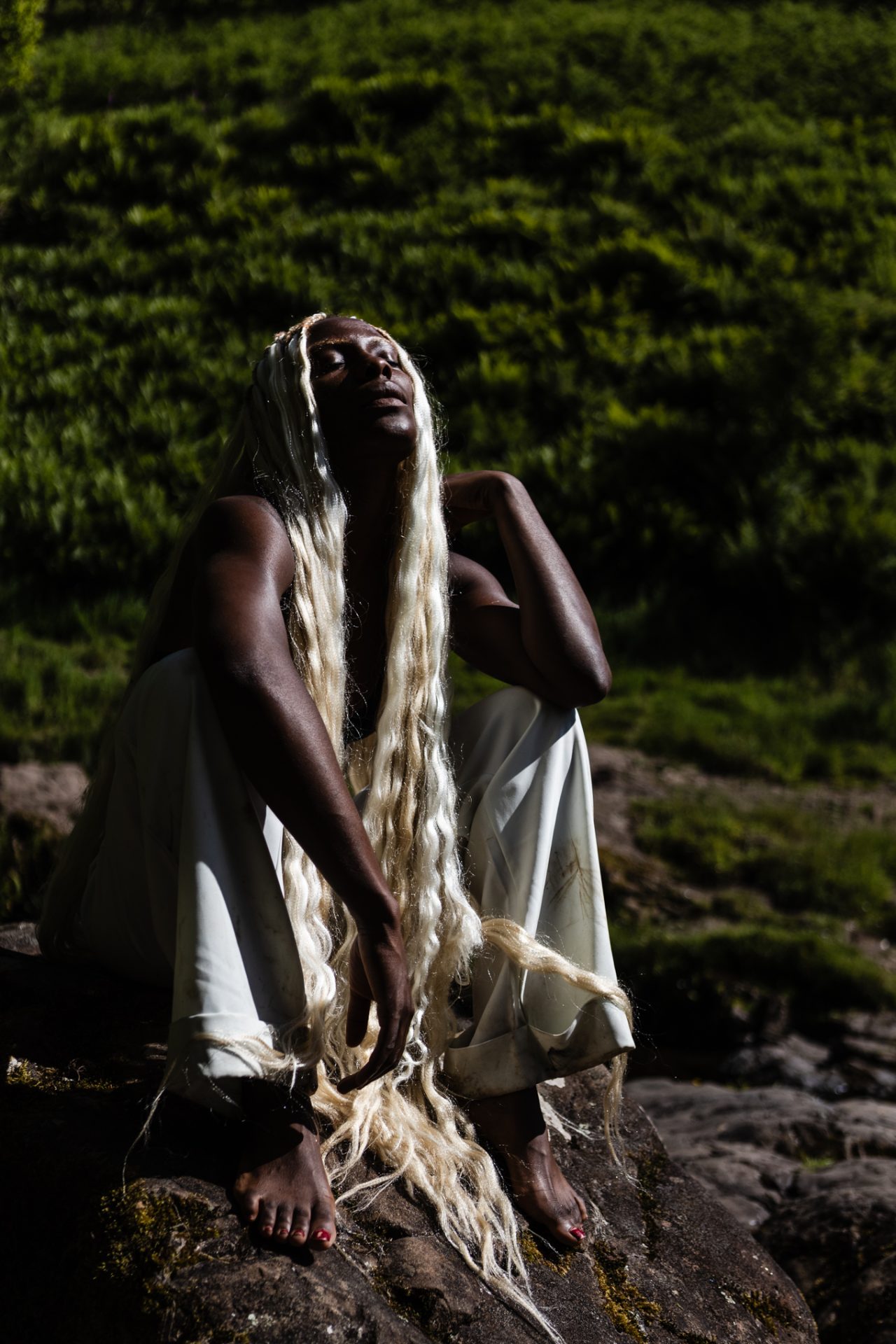 A Black woman with long, blonde hair sits on a rock. Hair eyes are closed, and she tilts her face towards the sun.