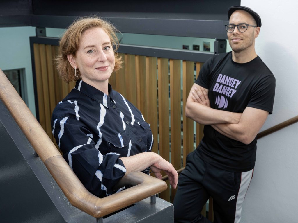 Two people stand in Dance Base's stairwell. One leans an elbow on the wooden banister, the other leans against the wall behind. They both smile into the camera.