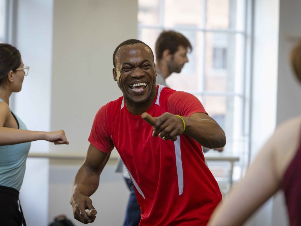 A smiling man in a bright red t-shirt points towards the camera.