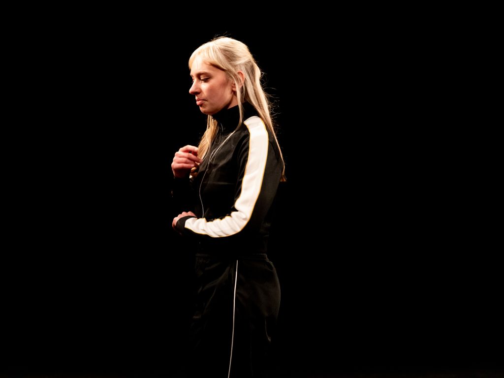 A dancer in a black tracksuit with a white stripe down the arm stands on a darkened stage. The are brightly lit, and their blonde hair stands out brightly against the dark.