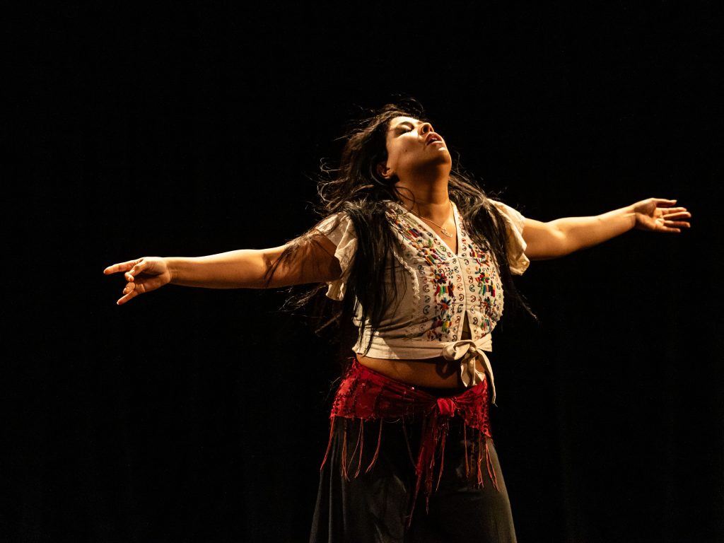 A dancer dressed in a white shirt embroidered with flowers stands with her head and arms thrown back.