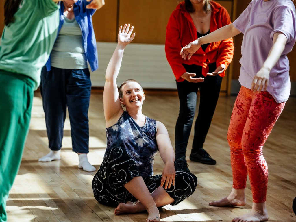 Community Company 12 June 2024. Photo Brian Hartley. A dancer, seated on the floor and surrounded by her fellow dancers, reaches her arm over her head, pointing up towards the roof of a sunlight-filled studio.