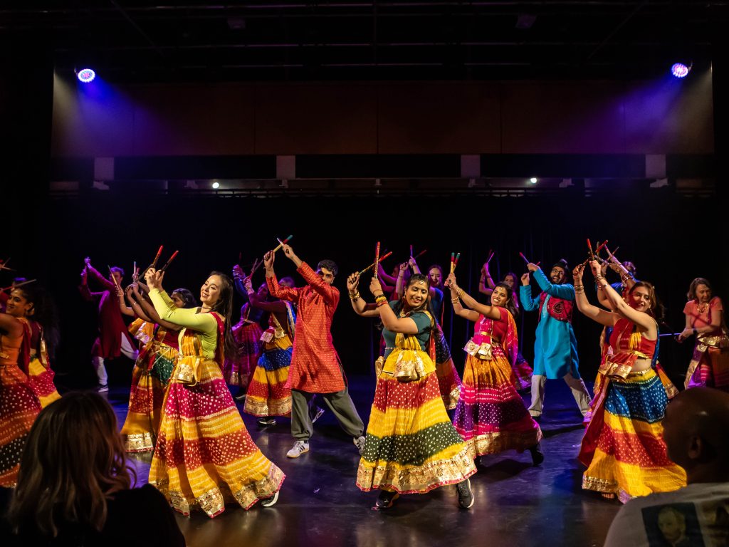 A group of smiling dancers in brightly coloured traditional Indian clothing dance on stage