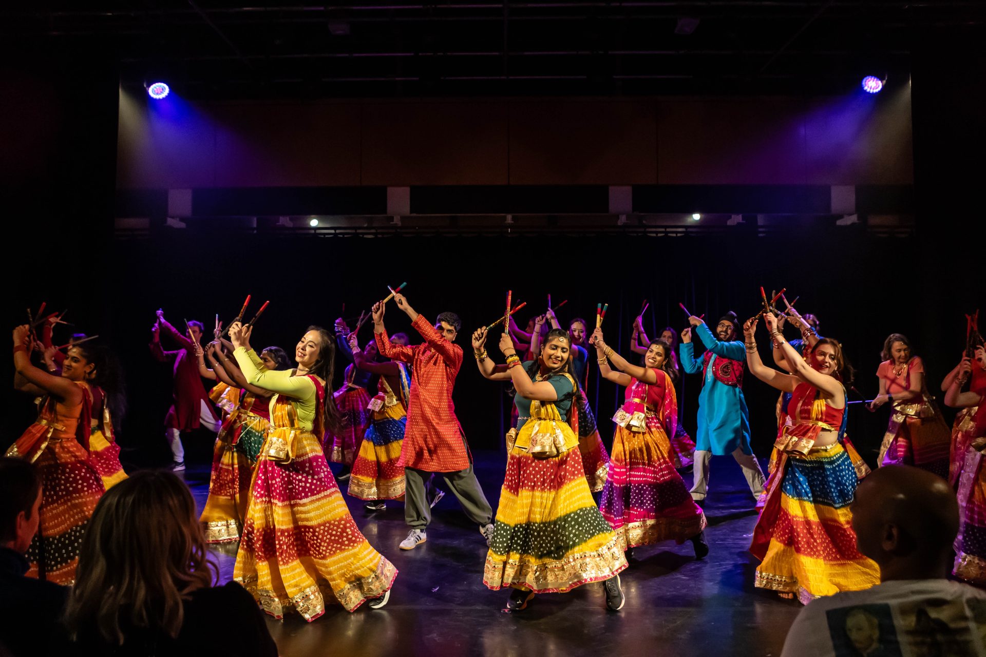 A group of smiling dancers in brightly coloured traditional Indian clothing dance on stage