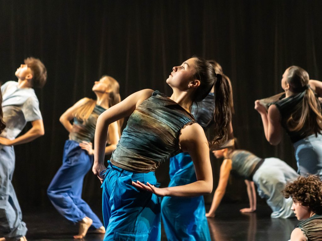 A group of young dancers in blue, caught in a pose leaning back with their hands on their hips