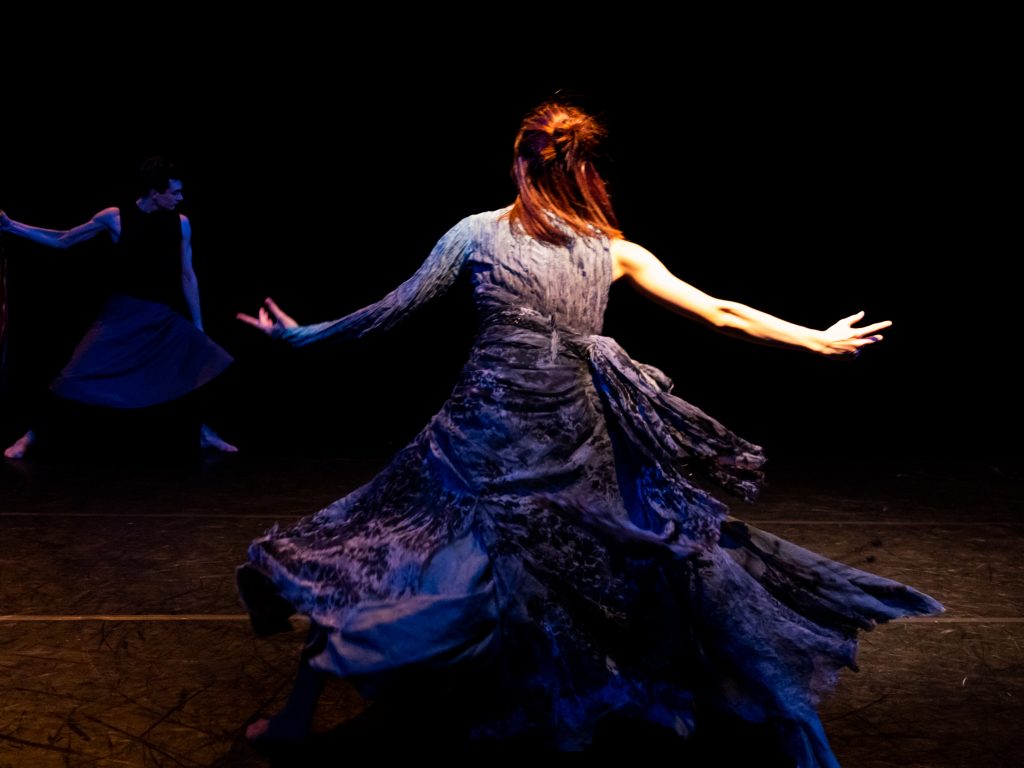 A dancer in a long, voluminous dress stands centre stage, back to the audience and arms outstretched. They are bathed in a blue light.
