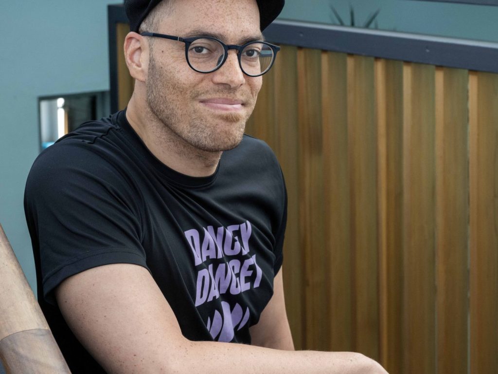 Tony Mills at Dance Base, September 2024. Image credit Neil Hanna Dance Base's Artistic Director Tony Mills stands in Dance Base's stairwell, wearing a black flat-cap and a t-shirt with the words: