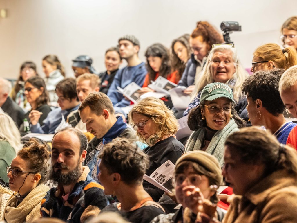 A seated audience in a theatre, smiling at one another and reading programmes.