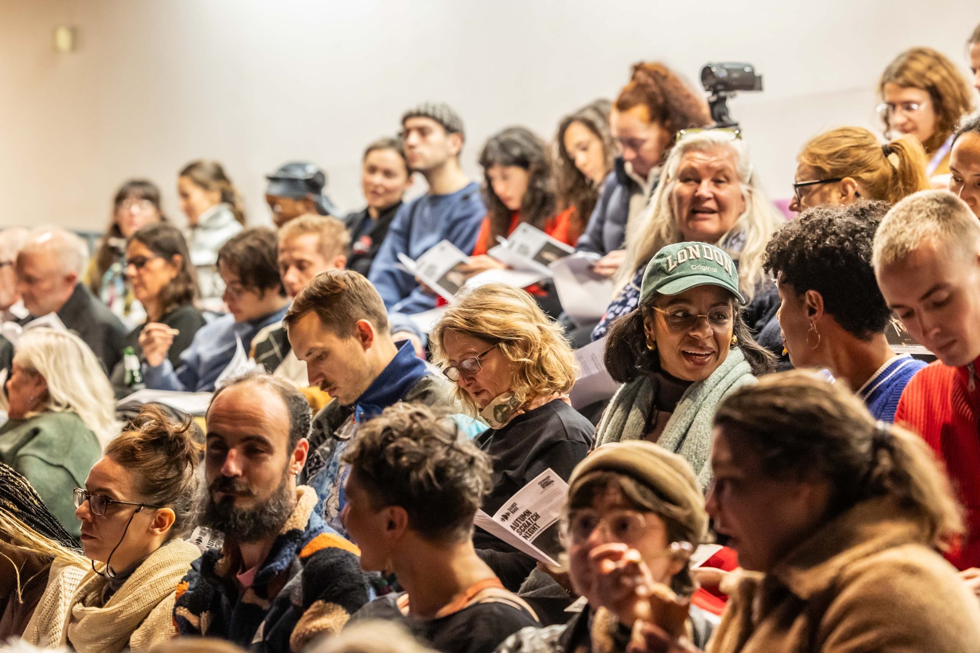 A seated audience in a theatre, smiling at one another and reading programmes.