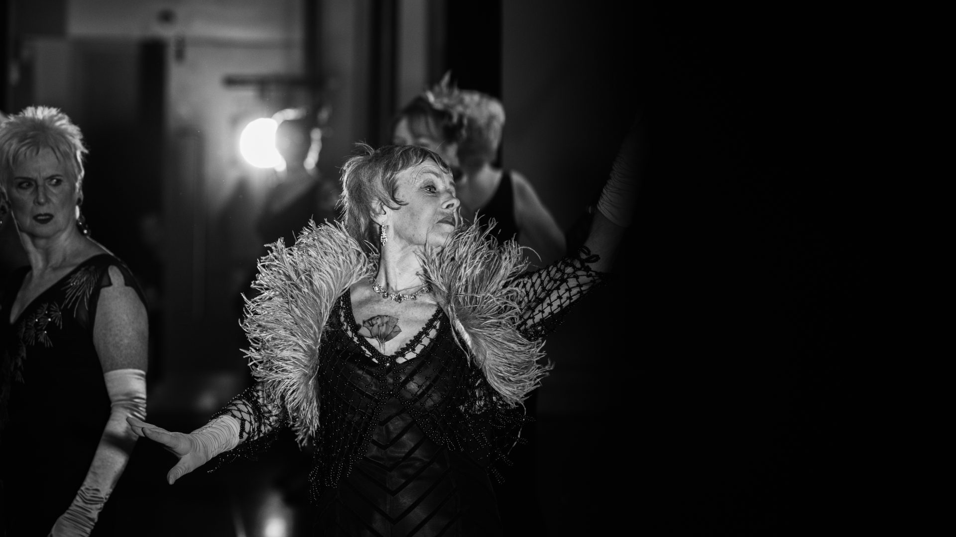 PRIME, Maureen in ‘Carry on Dancing’ by Steinvor Palsson, 2016. Photograph by Matt Austin. A black and white photograph of a woman in an evening dress and feather boa reaching her arms out wide.