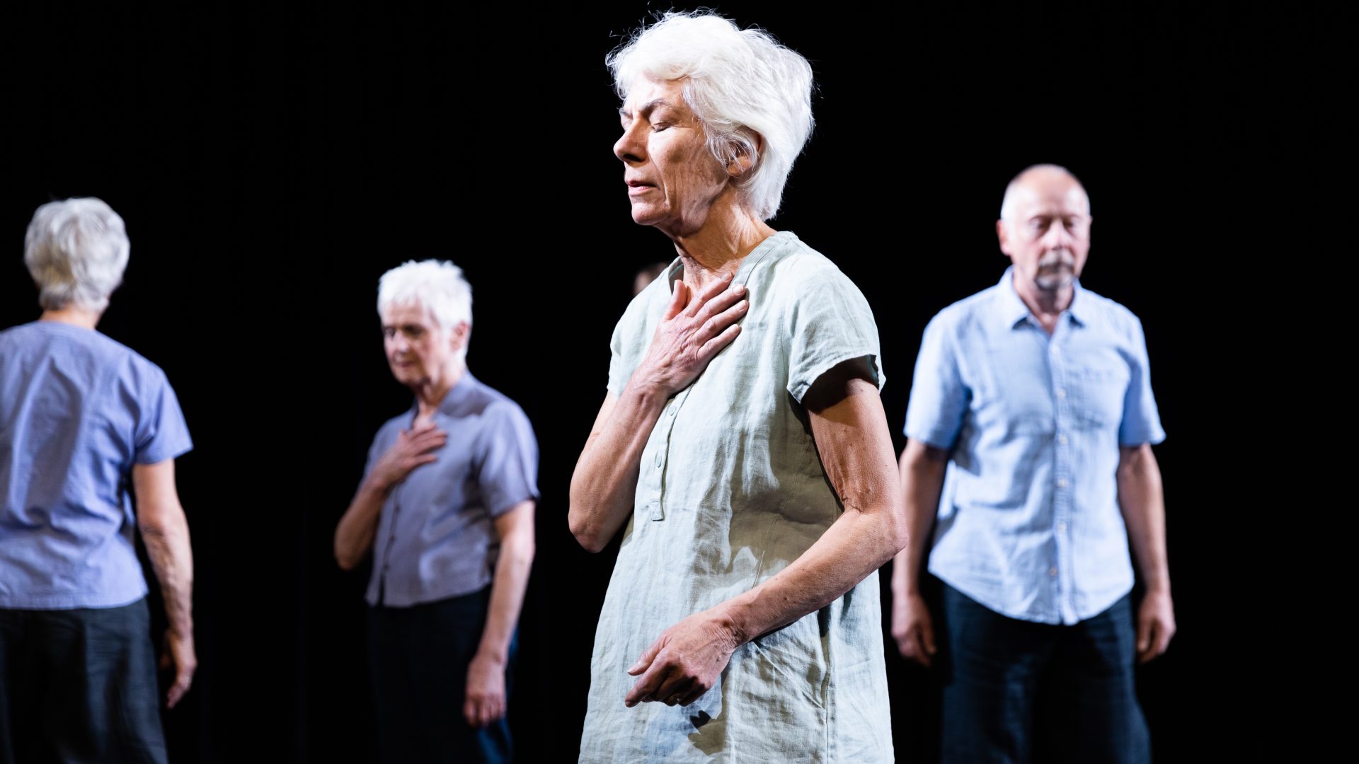 Amy Sinead 2024 Three dancers are pictured on stage. At the front of a picture, a woman is facing the diagonal and has one hand to her chest with her eyes closed.