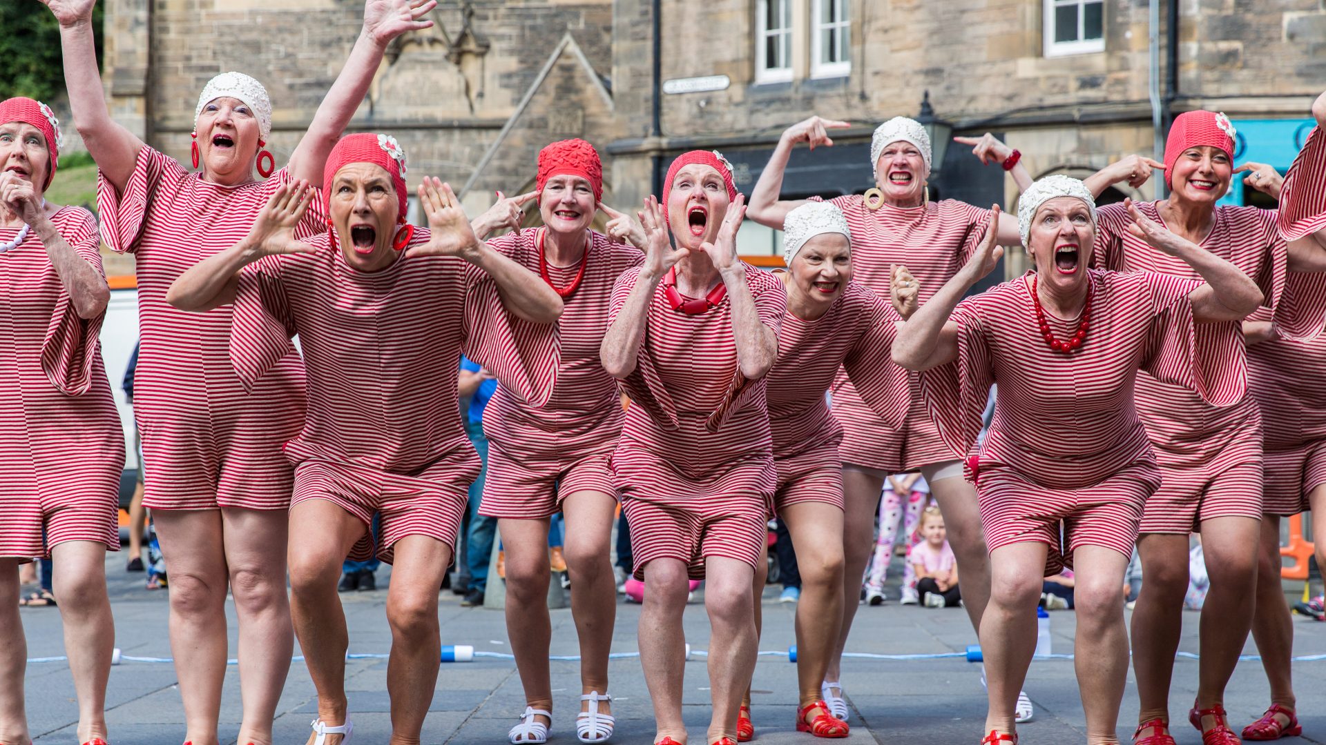 PRIME, ‘Bonnie and her Splashmob’ by Bridie Gane, 2018. Photograph by Amy Sinead. A group of dancers in red and white striped vintage swimming costumes that cover their thighs and upper arms. They are wearing swmming caps and swim shoes and posing with their hands near their heads or faces with mouths wide open.