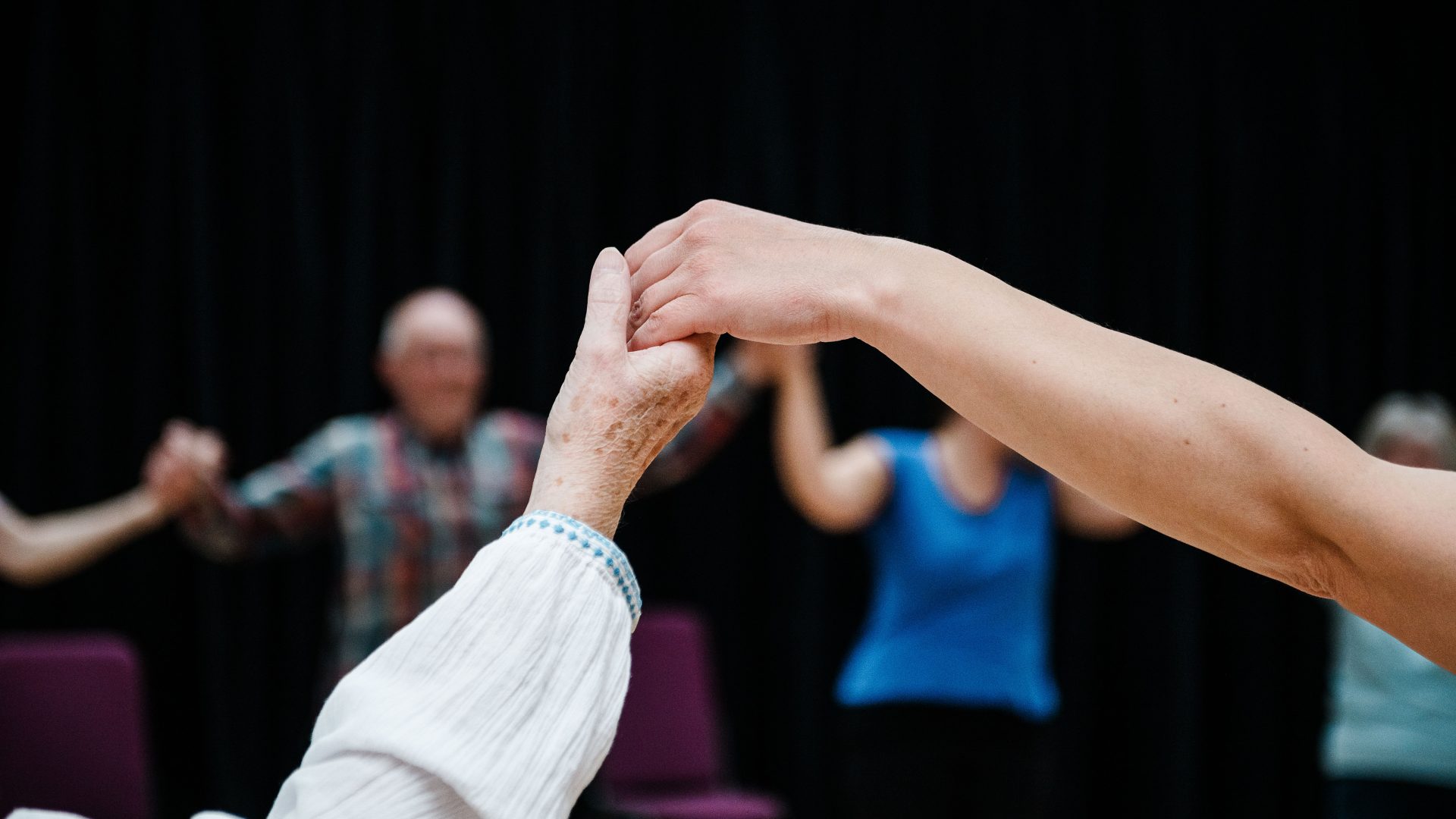 DB Parkinson's Class_12 June 24_Photo Brian Hartley_1754_hi A close up of two hands holding each other as part of a seated class. Blurry dancers can be seen in the background sitting in chairs and also holding hands to form a circle.