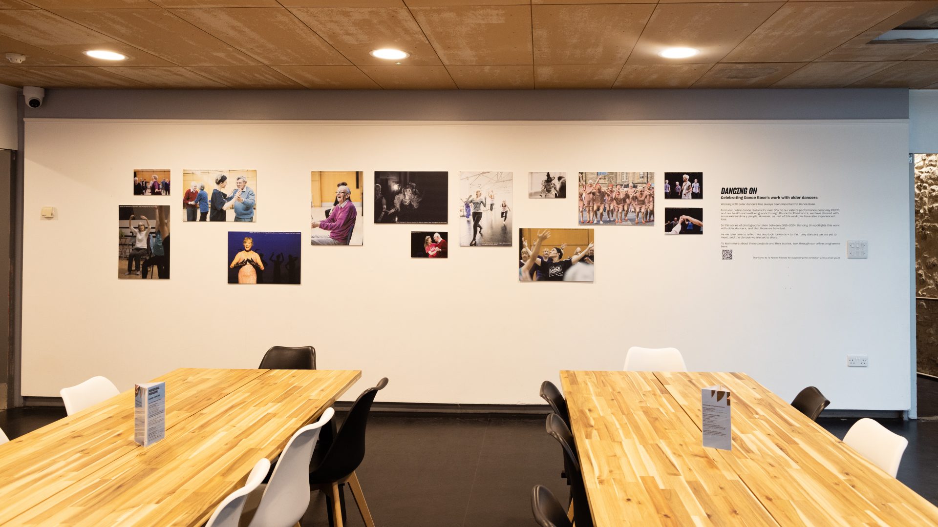 Dancing On Exhibition, Dance Base 2024-25. Image credit Stuart Armitt A front view of the exhibition hanging in Dance Base's upper foyer, taken from further away to show tables and chairs in the space.