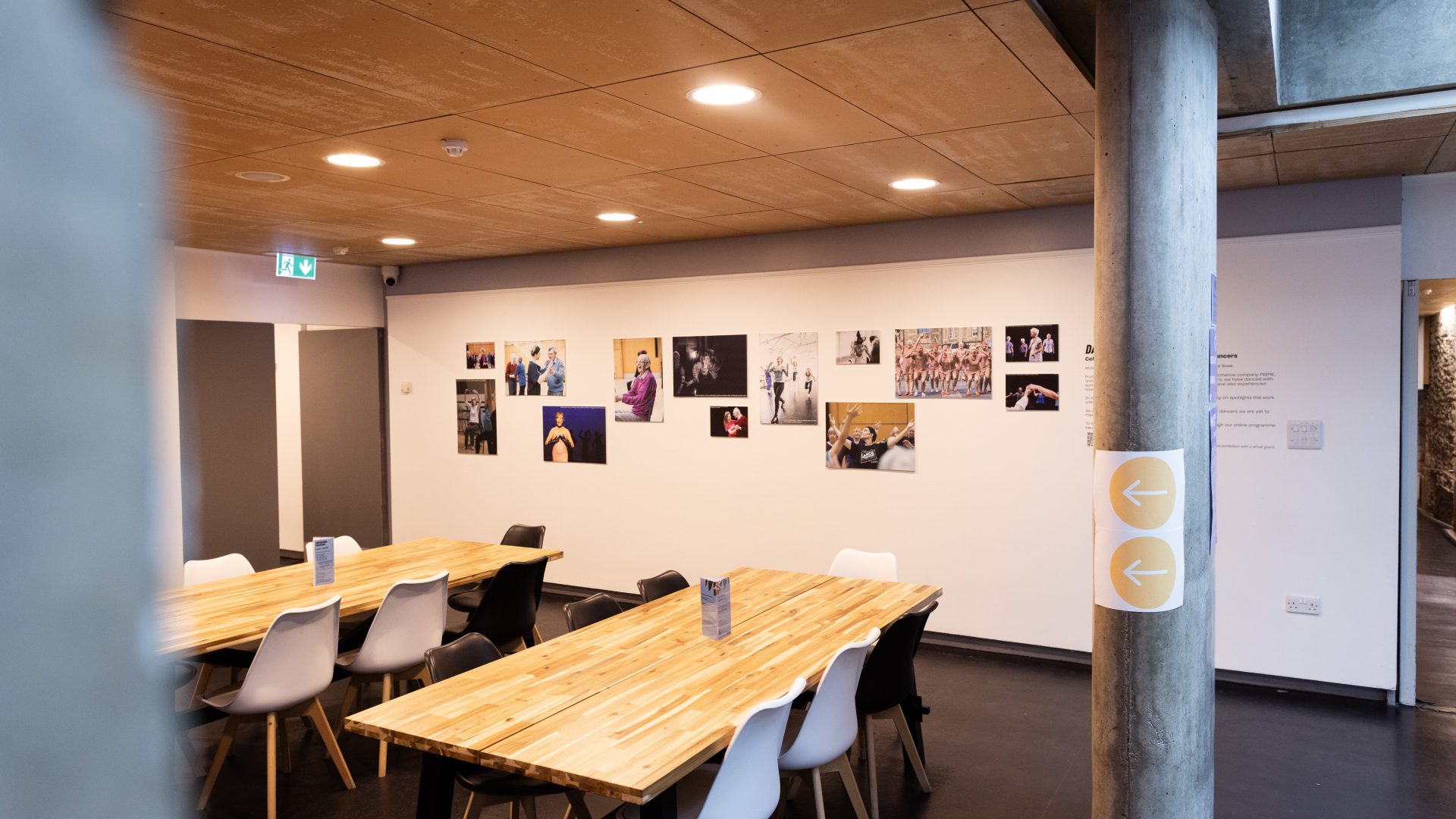 Dancing On Exhibition, Dance Base 2024-25. Image credit Stuart Armitt A side view of the exhibition hanging in Dance Base's upper foyer, taken from further away to show support pillars in the space as well as tables and chairs.