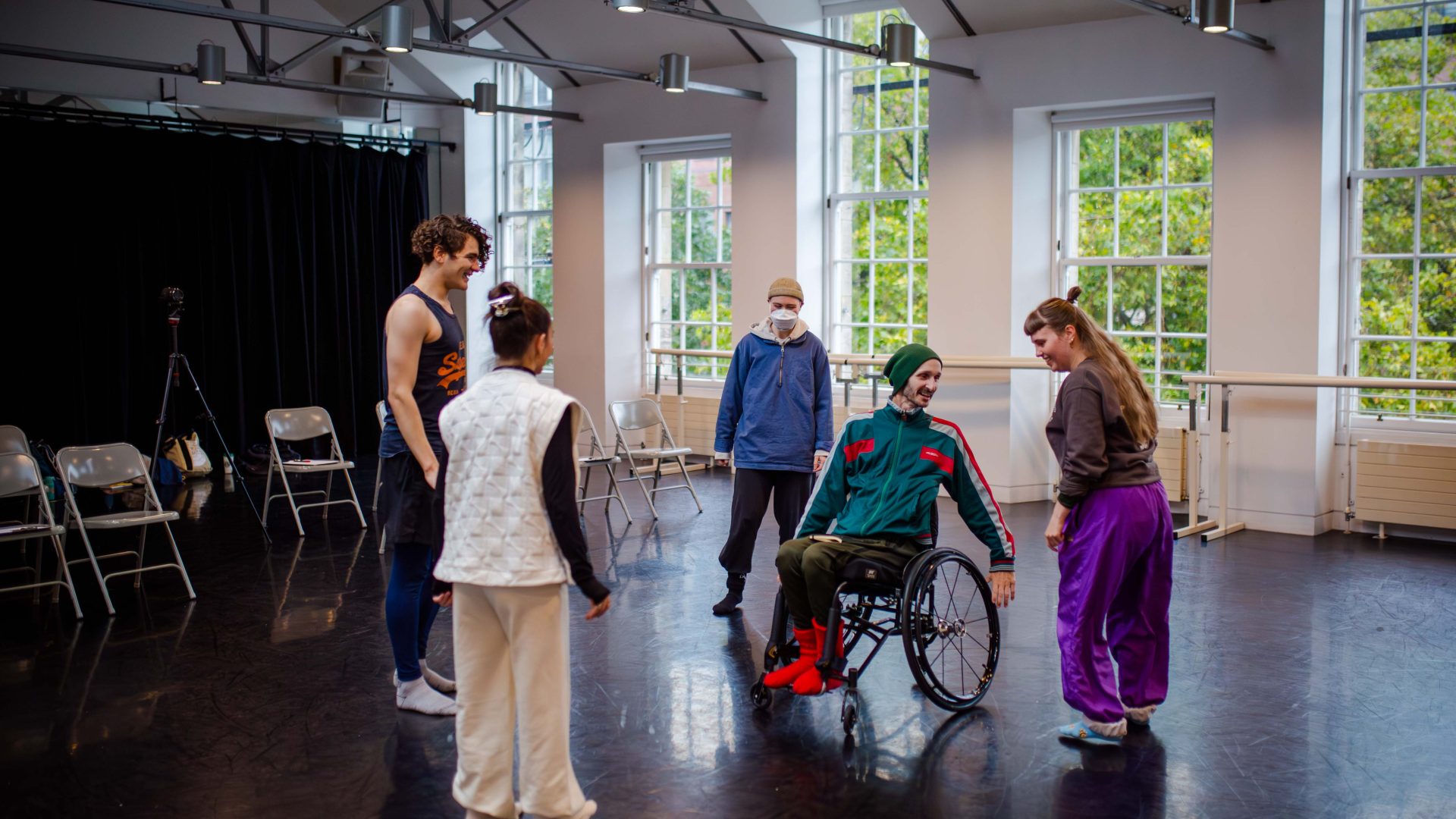CROPPED DEBS autumn residency 2025. Image credit: Paul Andrews A group of four young dancers stand in a bright studio with large windows, along with their mentor, who is using a wheelchair.