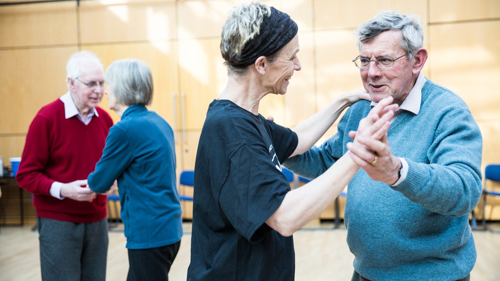 Dance for Parkinson’s class, 2019. Photograph by Amy Sinead. a man and woman are dancing in a bright dance studio. They are in a ballroom style hold at the front of the photo. In the background, there are two other people in a partner dance.