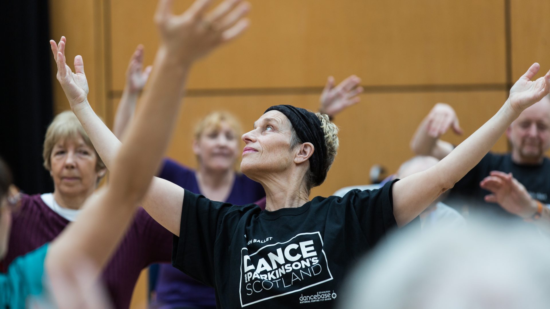Dance for Parkinson’s, Sophie, 2019. Photograph by Amy Sinead. A dance class close up. A woman in a Dance for Parkinson's t-shirt is in the process of raising her arms. She is surrounded by other dancers also raising their arms.