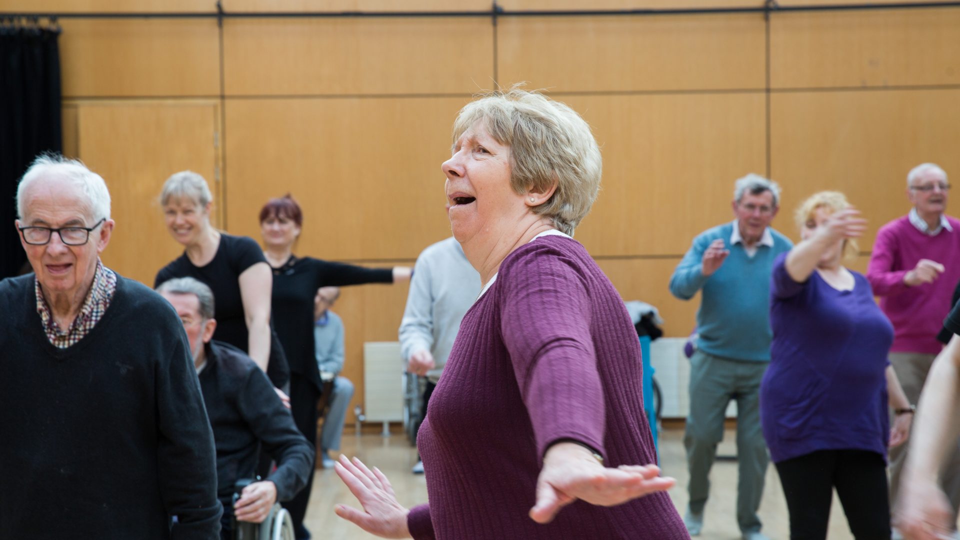 danceparkinsons-69 Amy Sinead A woman in a purple top is reaching her arms out to either side with a playful expression on her face. Other dancers are in the space moving their arms in different directions.