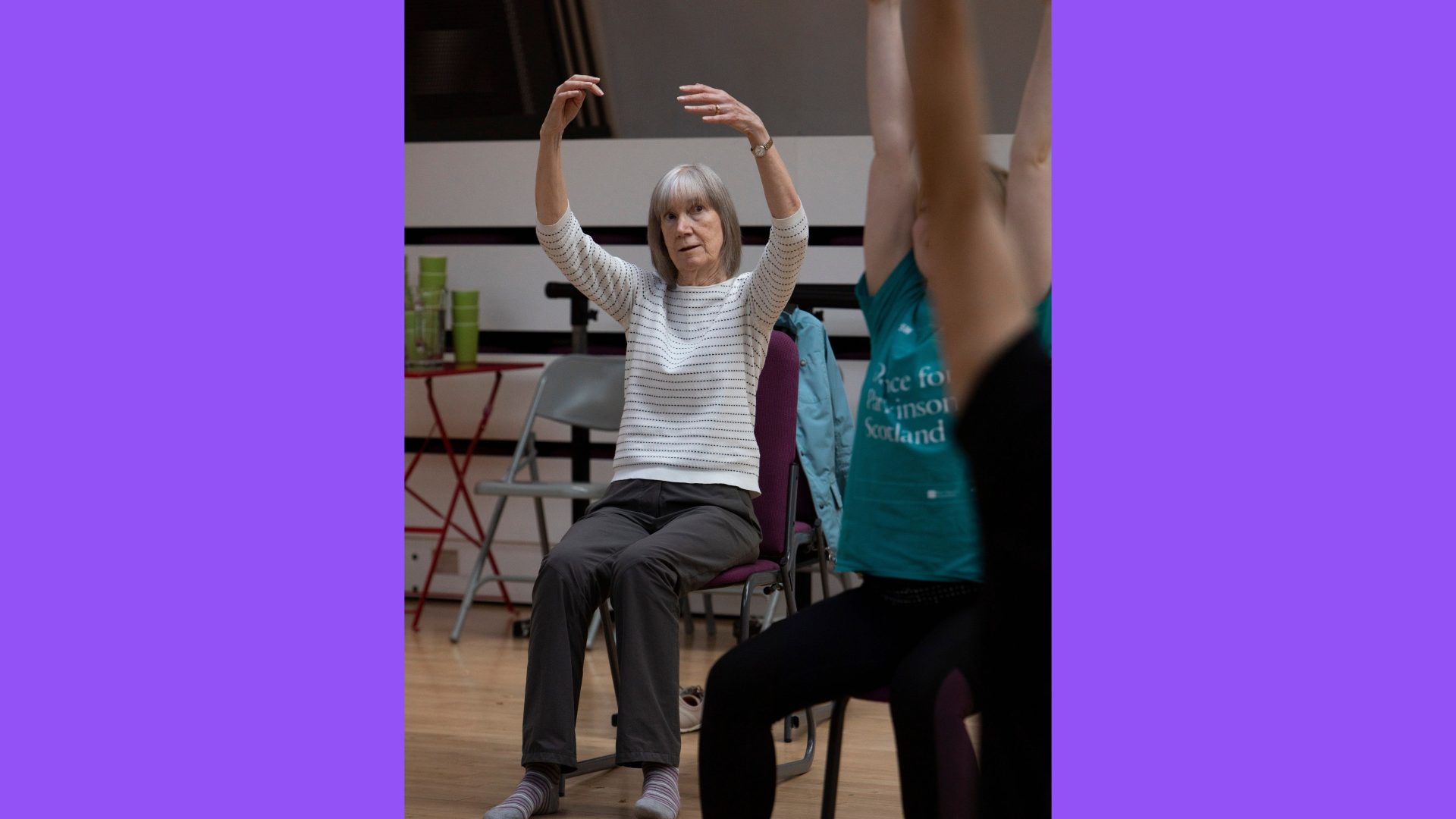 Dance for Parkinson’s, Gwynneth, 2022. Photograph by Amy Sinead. A close up of a dance class. A woman in a white top is seated in a purple chair with her arms raised above her head.