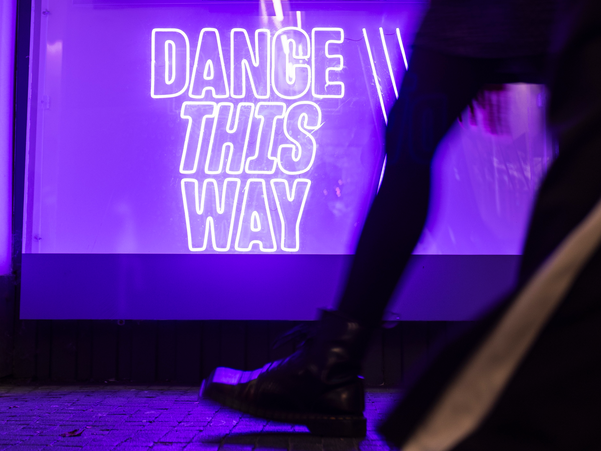 Dance Base Pend, 2025. Image Credit Eoin Carey. A close up photograph of two sets of legs walking in front of a video of Artistic Director Tony Mills and a purple neon that says 'Dance This Way'.