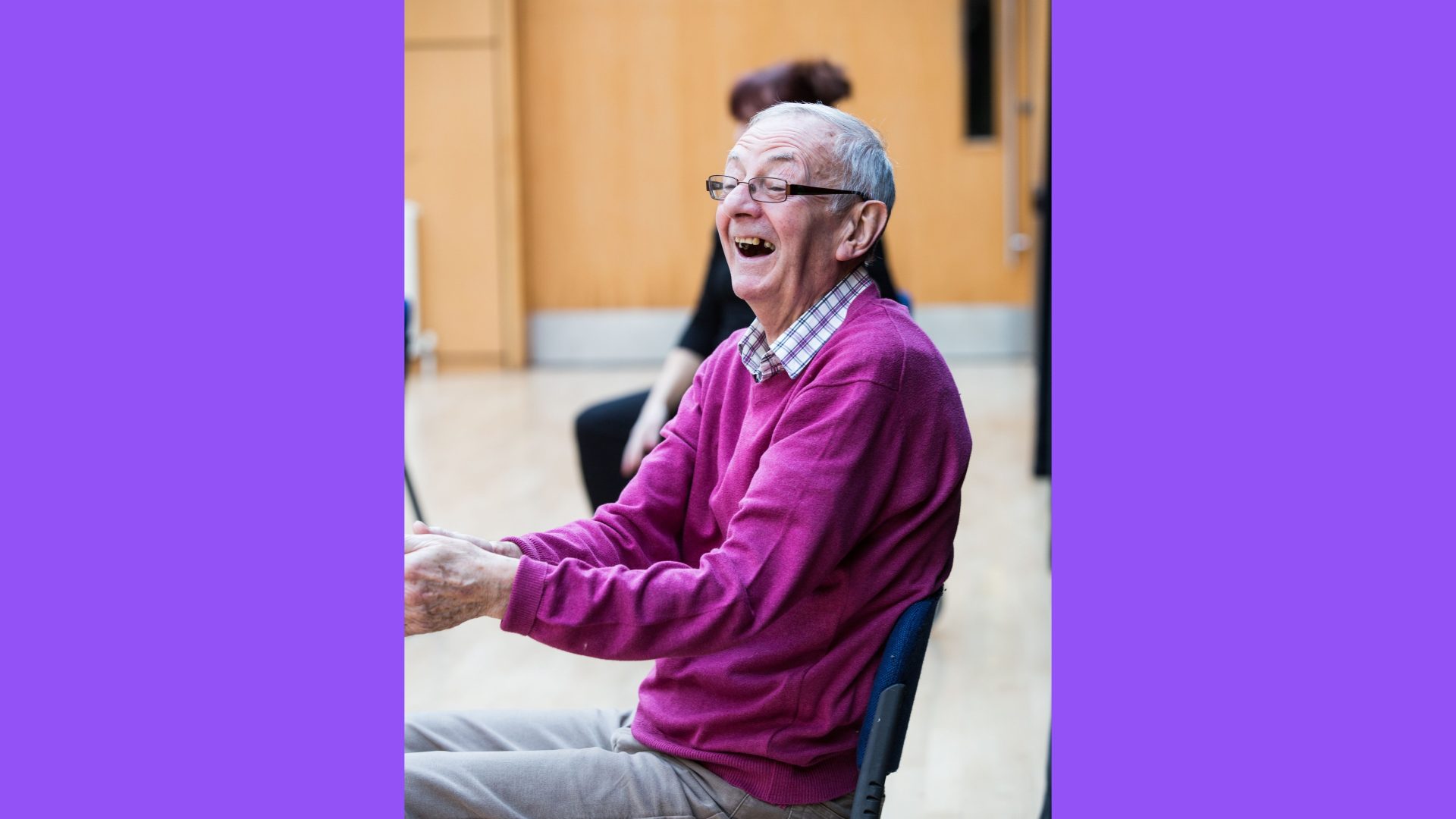 Dance for Parkinson’s, Reg, 2019. Photograph by Amy Sinead. A man in a bright purple jumper and checked button up shirt is laughing while clapping his hands.
