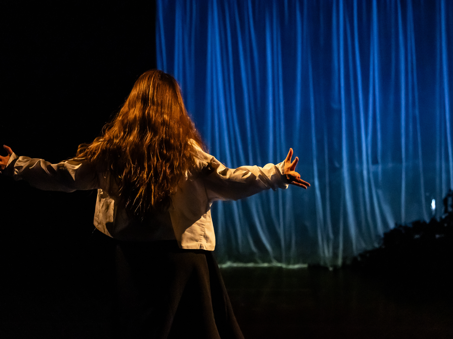 CROPPED Damara Velázquez Pastrana in Dance Base’s Autumn Scratch Night 2024. Image credit Sally Jubb. A dancer faces a blue curtain at the back of the stage, arms outstretched.