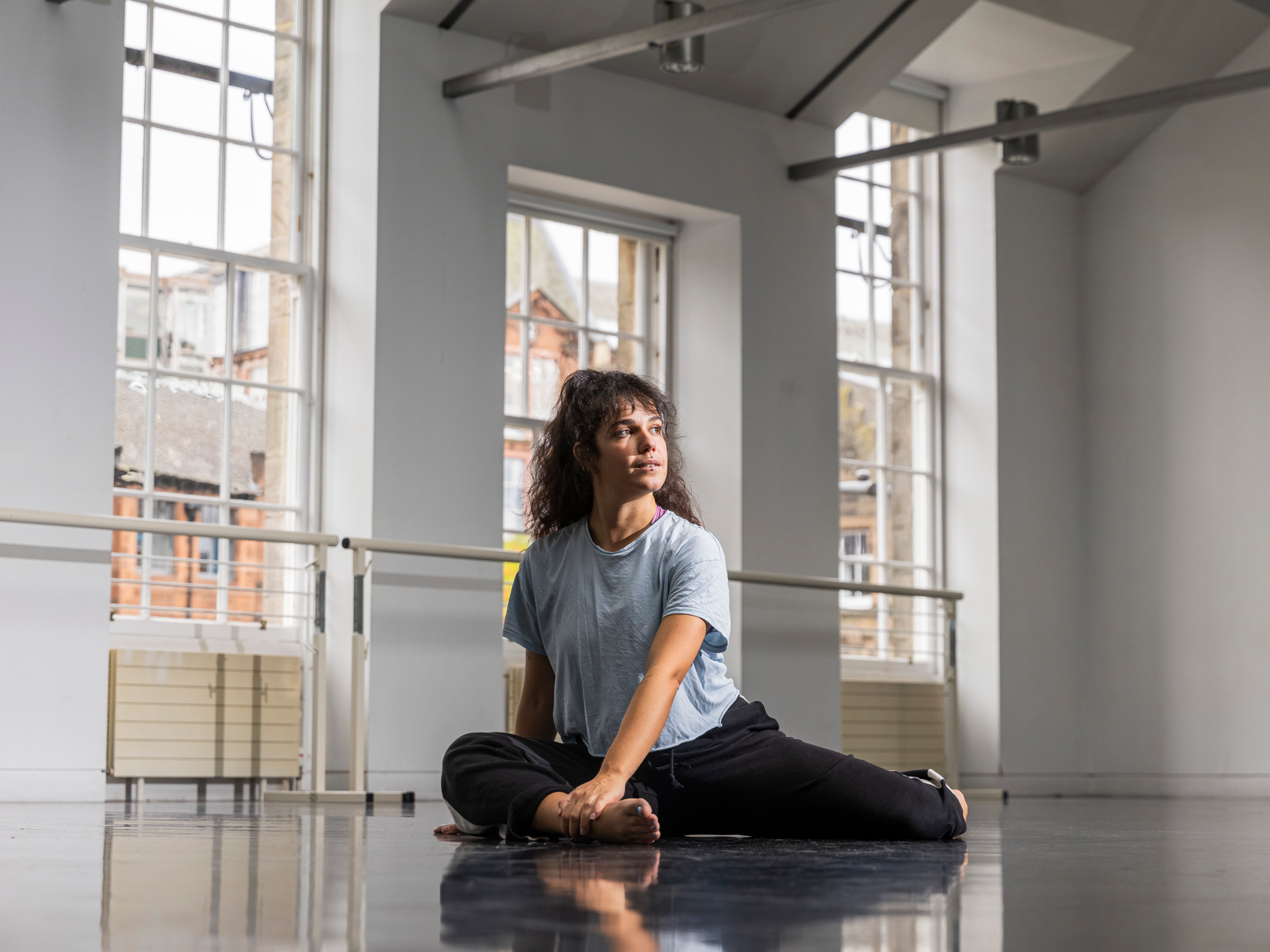 CROPPED Alex Tsiapi in Dance Base studio 2. Image credit Eoin Carey A dancer sits on the floor before the large windows of a bright dance studio.