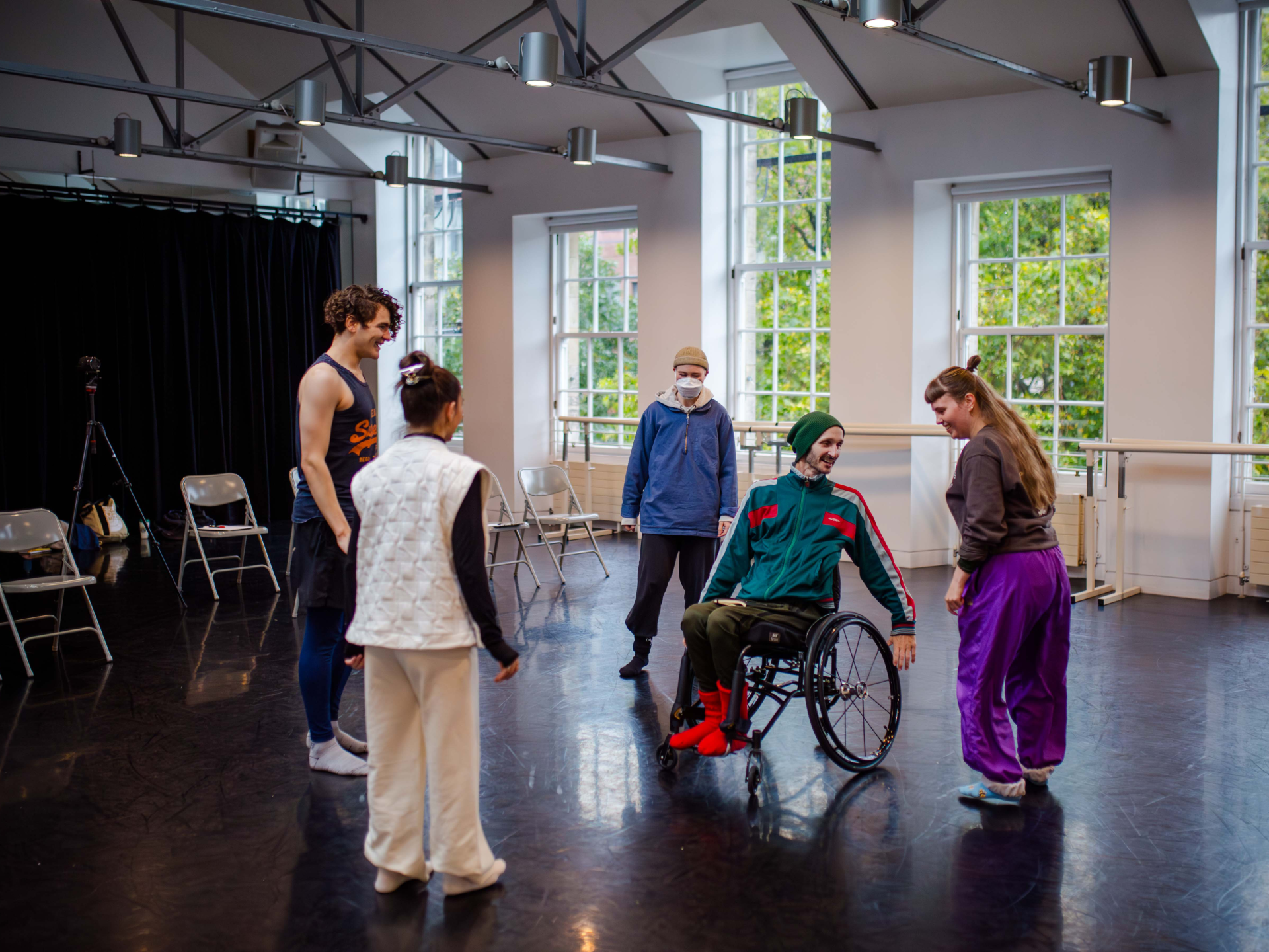 Cropped for ‘About’ DEBS autumn residency 2025. Image credit: Paul Andrews A group of four young dancers stand in a bright studio with large windows, along with their mentor, who is using a wheelchair.