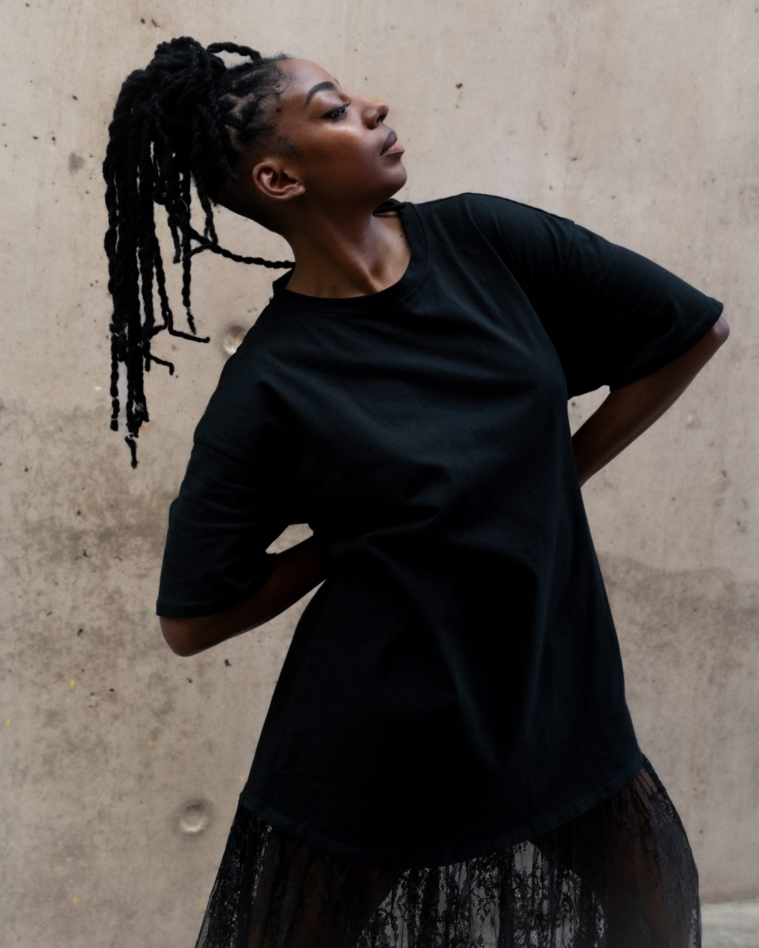Mele Broomes. Image credit Izzy Leach. A photograph of a dancer all in black, posing against a rough stone wall.