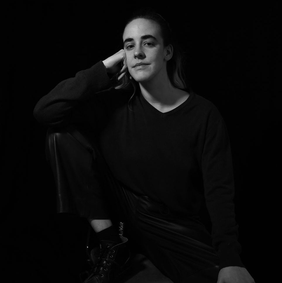 Kirstin Halliday headshot. Image credit: Phyllis Christopher. A black and white headshot of a dancer, sitting with one foot up on a chair.