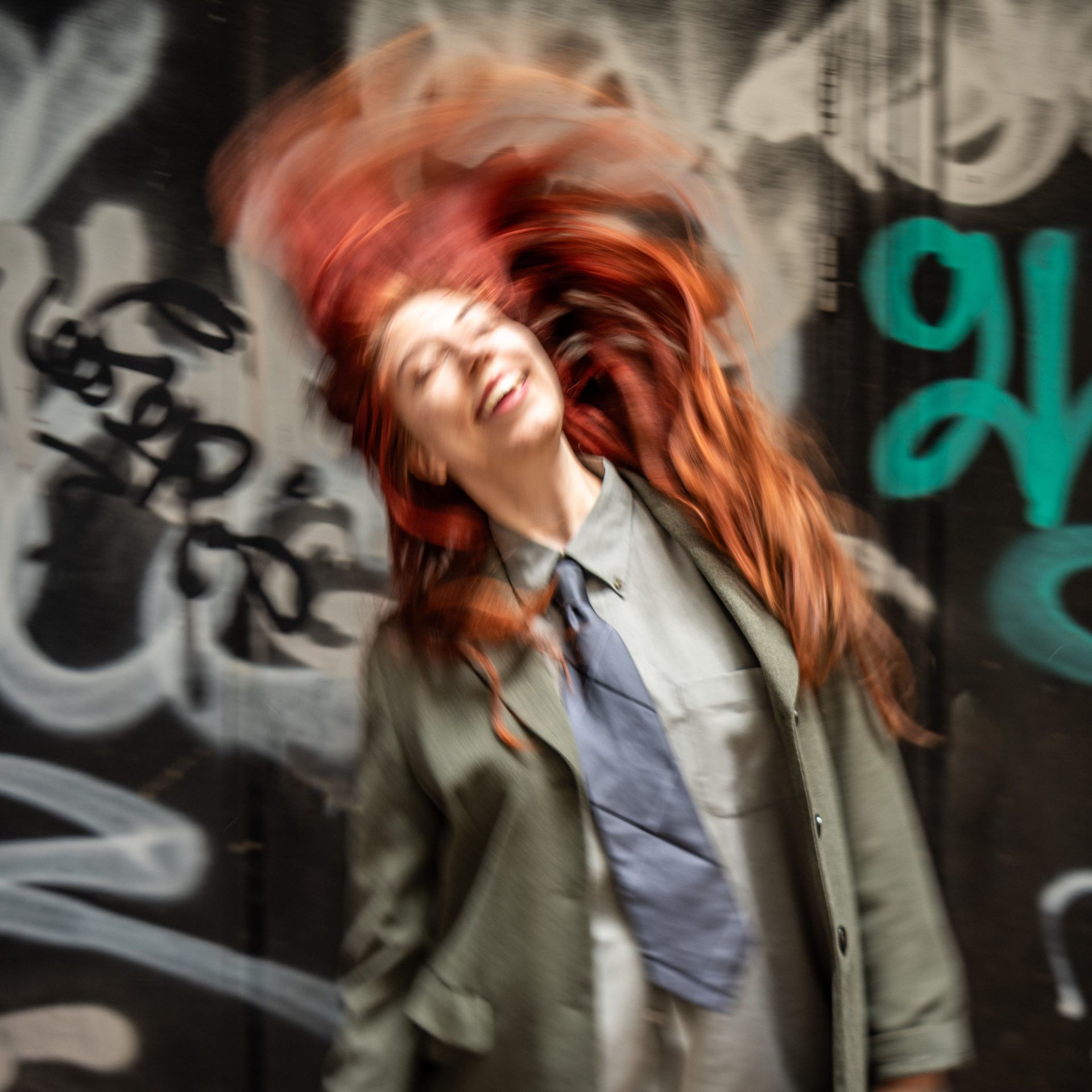 Evellina Tiger A dancer with red hair stands in front of a graffiti'd wall, flicking their hair over their shoulder.