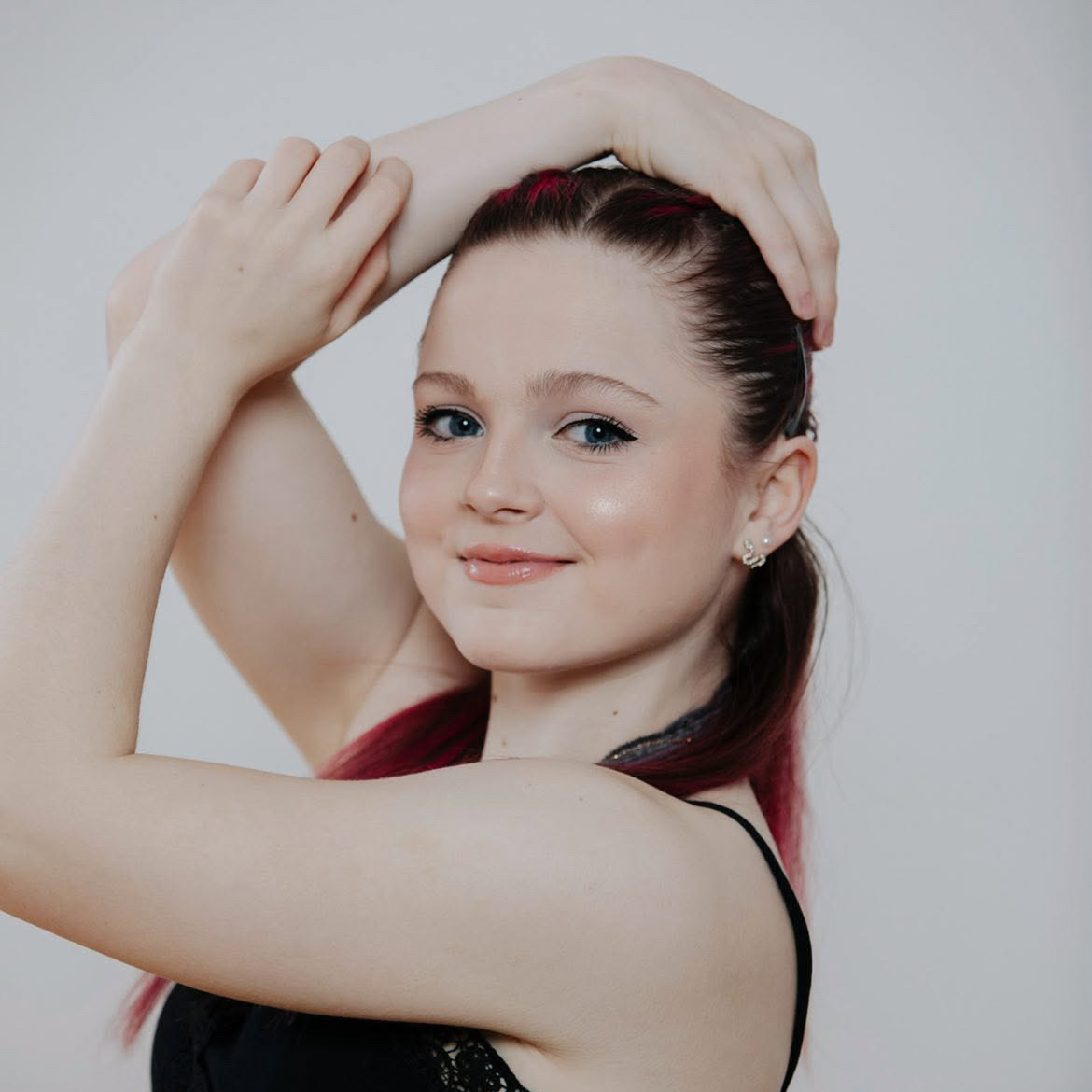 Sophie Morrison A headshot of a dancer with their arms over their head, looking towards the camera